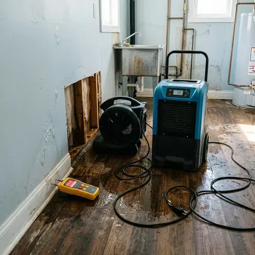 Air mover, dehumidifier and moisture meter drying a wet wood floor beside an opened wall