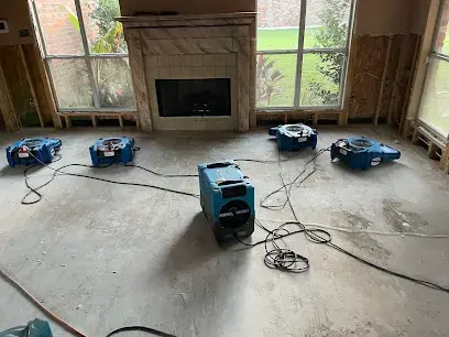 Air movers and a dehumidifier drying a stripped floor in front of a fireplace